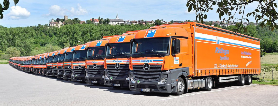 You can see a huge tarpaulin low loader with both tarpaulins completely open. On the loading area is a huge machine. You look at the truck from the side. In the background two other trucks are partially visible.