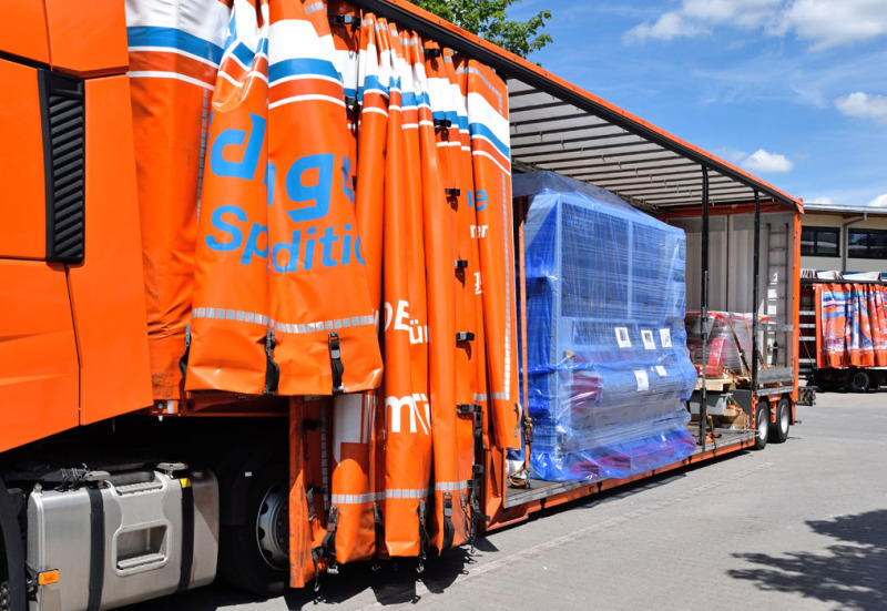 You can see from the front on a tarpaulin low loader, from the opened tarpaulin. On it stands a blue machine, which is shrink-wrapped in foil. In the background you can see another truck.