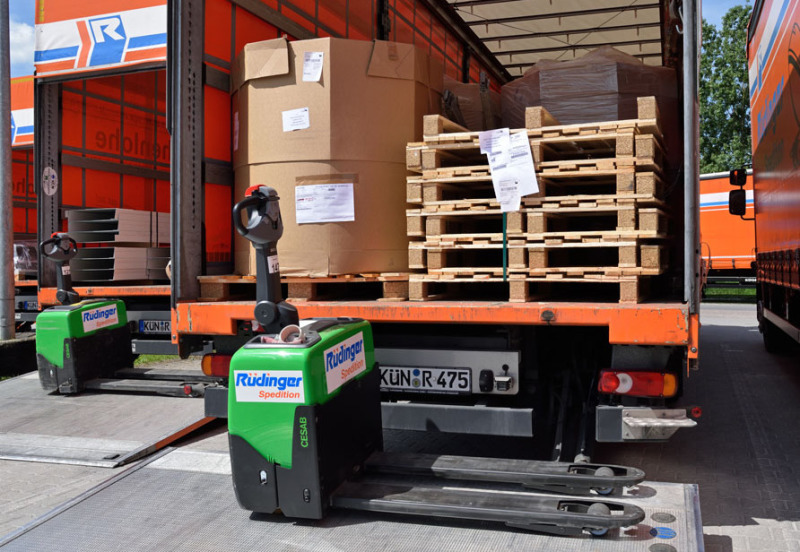 Two 7.5 ton trucks are photographed from the rear and open. In front of both is a black/green electric pallet truck. Both loading platforms are loaded with goods. In the right one there are pallets and two cartons.