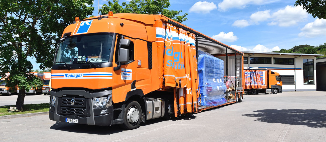 You look at a huge tarpaulin low loader from the front and from the side. The side curtain is open and on the loading area is a huge, blue, shrink-wrapped machine. In the background there is another truck and you can see the administration building.