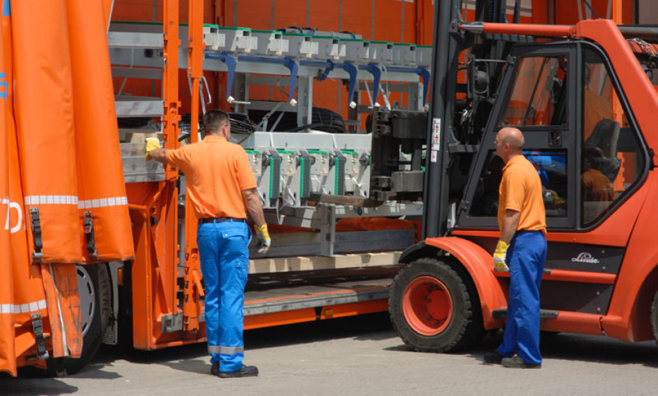 Two emplyees with orange and blue clothes care about a big machine, which a forklift placed into a trailer. On the right side there is the forklift.