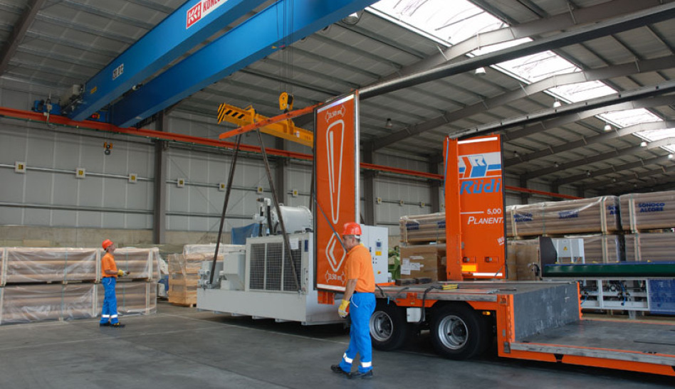 Two emplyees in blue and orange chlothes and a helmet in orange work on a big gray machine, to place it on the floor of a completely open trailer floor in a warehouse.
