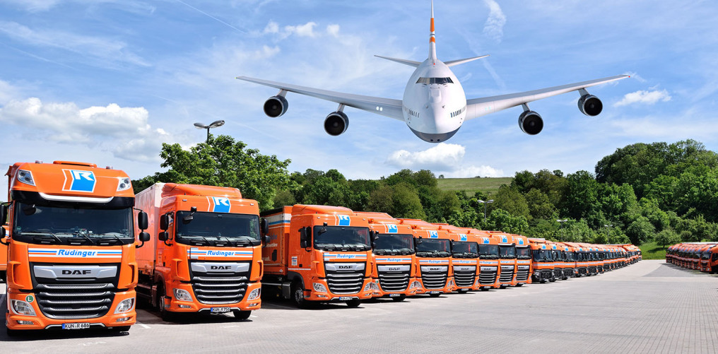 Two rows of huge trucks is arranged to perfection. There is an airplane flying low towards the photographer. Of course it is a photo montage. In the background there are trees and behind is a meadow on a hill.