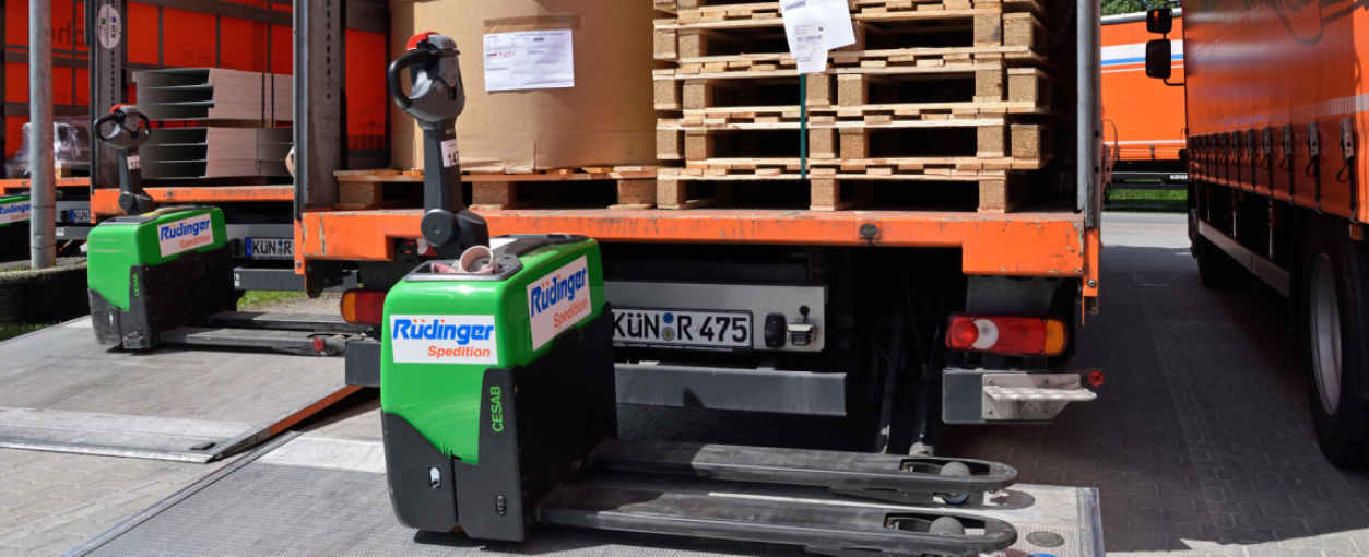 Two electric pallet trucks stand in front of the rear openings of two 7.5-ton trucks. There are goods on both loading platforms. On the left are two cartons and several pallets.