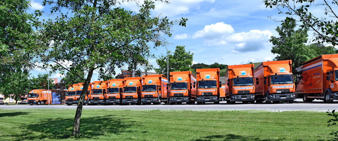 Two electric pallet trucks stand in front of the rear openings of two 7.5-ton trucks. There are goods on both loading platforms. On the right are two cartons and several pallets.