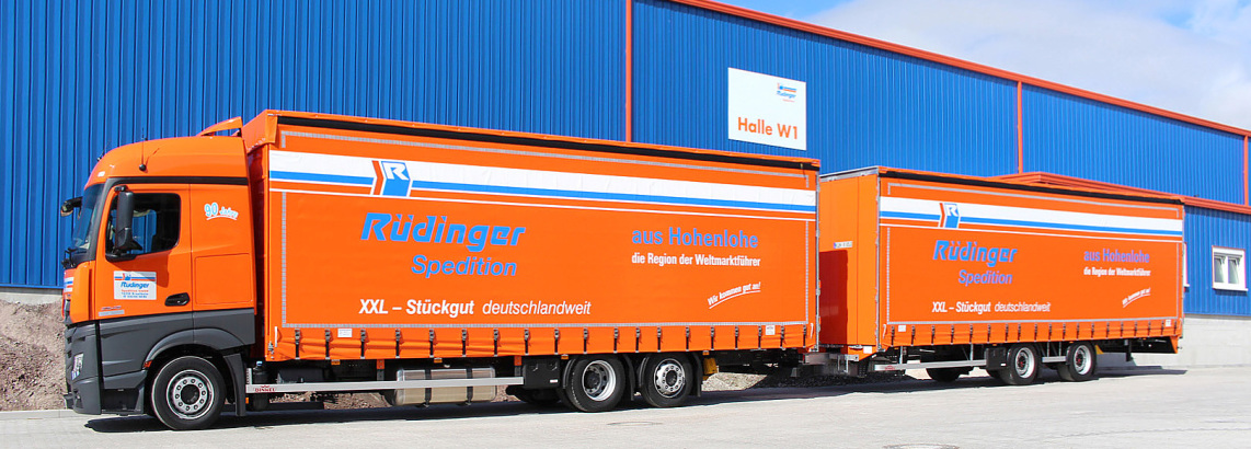 An extra-long truck, called a megaliner in Germany, is parked in front of a new blue warehouse. You can also see orange parts of the hall. The sky is blue.