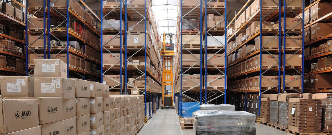 An extra-long truck, called a mega liner in Germany, is parked in front of a new blue warehouse. You can also see also orange parts. The sky is blue.