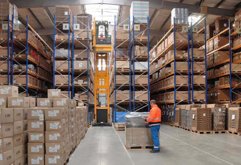 A new warehouse from the inside. You can see many rows of shelves, which are almost completely full. In the middle, an employee stands on a transport cart with goods.