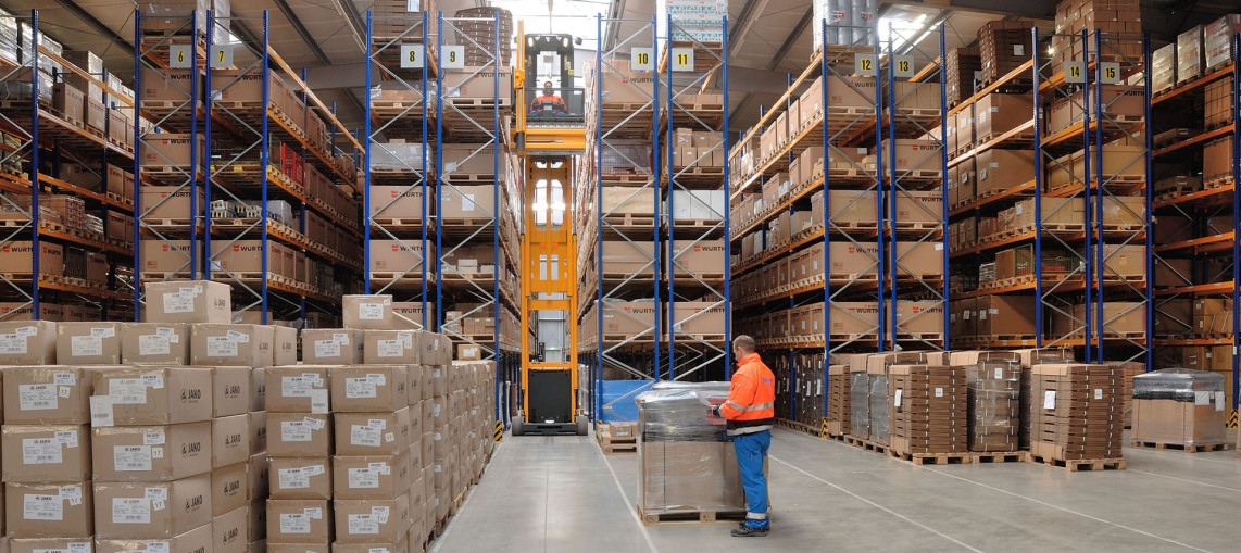 You look at the front of bulging shelves in a newly built warehouse. In the foreground, an employee is checking something on a pallet of goods.