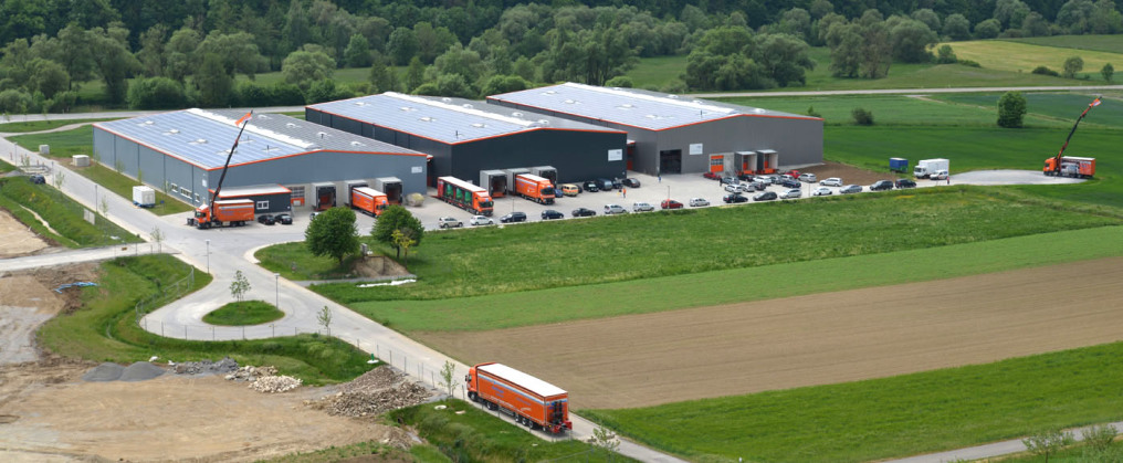 Photographed from above from the nearby vineyards, one looks down on three new-build warehouses of the logistics company in the midst of green fields and forest. On the left in front is a turning plate and on the photo you can see several tarpaulin low lo