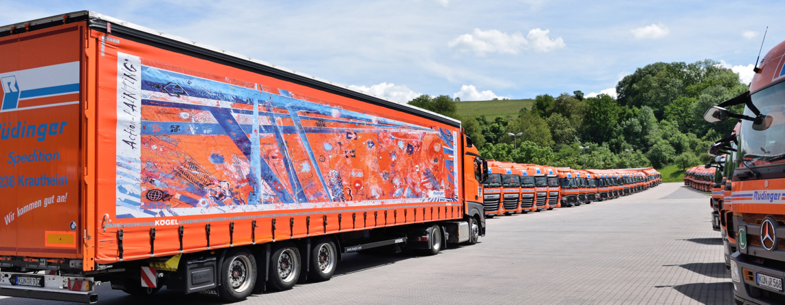Two rows of trucks are lined up side by side, so you can barely see the last trucks. Behind them are trees, meadow and a hill. In the center left, a special truck drives away from the viewer: the tarp is a work of art in orange and blue.
