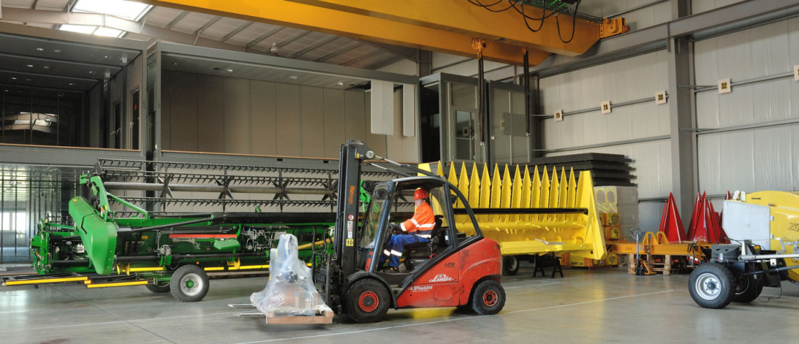 In the rear area of a newly built warehouse you can see two agricultural vehicles. One light green and one bright yellow. In front of them, an employee is driving a forklift truck and has a small packaged good on a pallet on the fork.