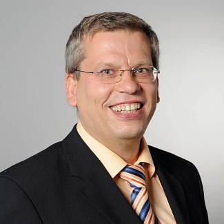 Roland Rüdinger is smiling heartily to the viewer. He has a black jacket on and an orange tie.