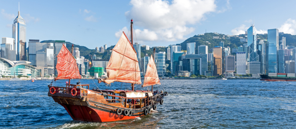 A red and brown junk sails in front of the skyline of Hong Kong, behind it you can see green mountains and above it a blue sky with white clouds.