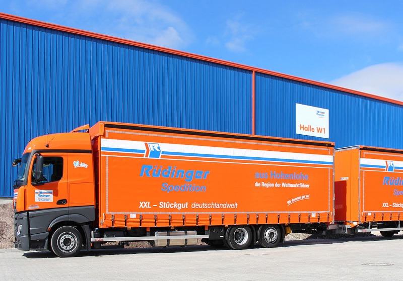 An orange tarpaulin low loader stands in front of a new warehouse in blue with orange parts. Only half of the trailer is visible.