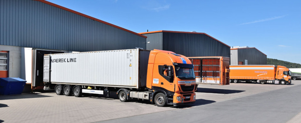 A truck with a container chassis and a silver container stands in front of a loading ramp of three warehouses. In the background you can see two more trucks. The sky is blue, the sun shines on the scenery.