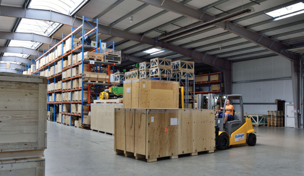 You look partly into the depth of a new warehouse, inside. All shelves are full of goods. At the front, an employee sits on a forklift and transports a very large wooden crate.