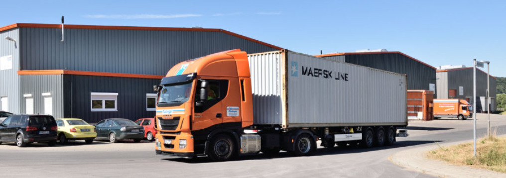 A truck with a large container leaves the freight forwarding company. In the background you can see three new warehouses and two more trucks. The sky is cloudless blue. A few cars are parked in front on the left.