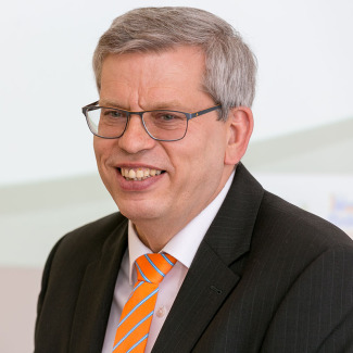 Roland Rüdinger smiles warmly at the camera. He is wearing a black jacket and a blue-orange tie.