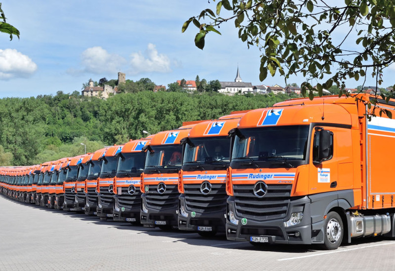 A long row of neatly parked trucks. Behind the row you can see a forest area and above it the "skyline" of Krautheim, Germany: a castle on the left, a church and houses on the right.