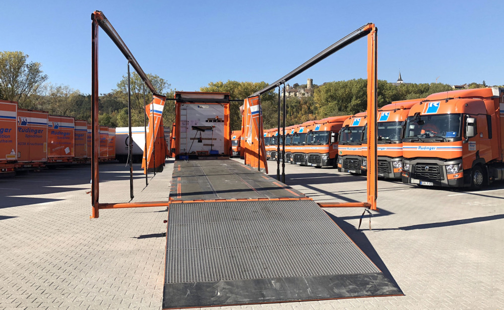 A tarpaulin low loader with the tarpaulin curtains completely open. The orange frame that remains is fully extended in width. A full-width ramp is lowered at the rear.