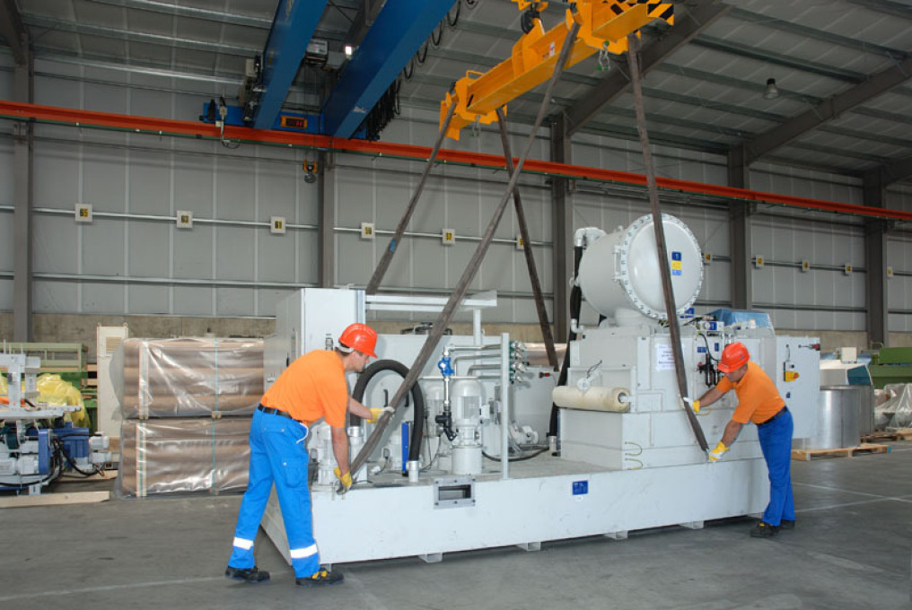 In a warehouse, two employees in blue/orange clothing with an orange helmet are working on the holding devices of a crane. A large gray machine is to be transported.