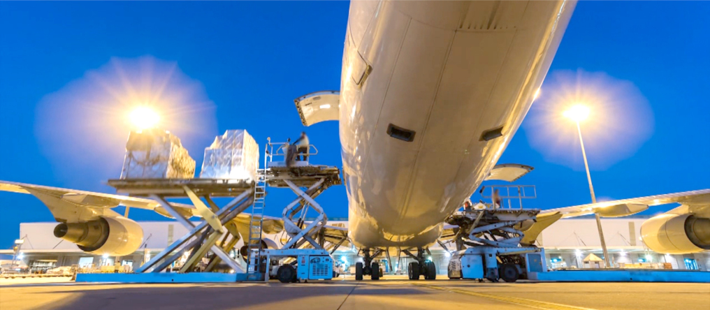 The loading of an airplane in the dusk. You look under the plane through to the back. On the left a loading platform is lifted up and there is goods on it. two bright lamps are on the right and left.