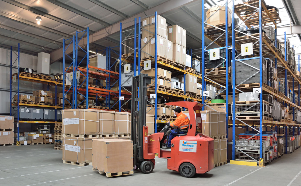 In front of many shelves in a newly built warehouse stands an angular forklift on which an employee is sitting. He has loaded a package onto a pallet. The shelves behind it are filled to the brim with a wide variety of goods.