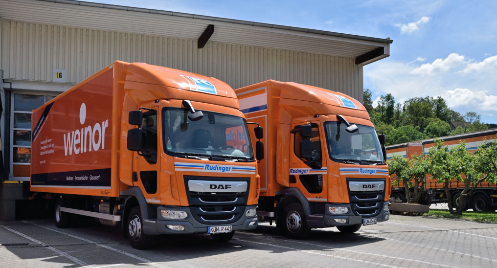 Two small, 7.5-ton trucks painted orange stand in front of a ramp of a warehouse. You look at them diagonally from the front. The one on the left advertises the Weinor company, with which the logistics company has been working for a long time.