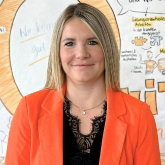 Sansra Platzgummer is warmly smiling to the viewer. She is standing in front of an art work. She wears a white t-shirt and a beige jacket.