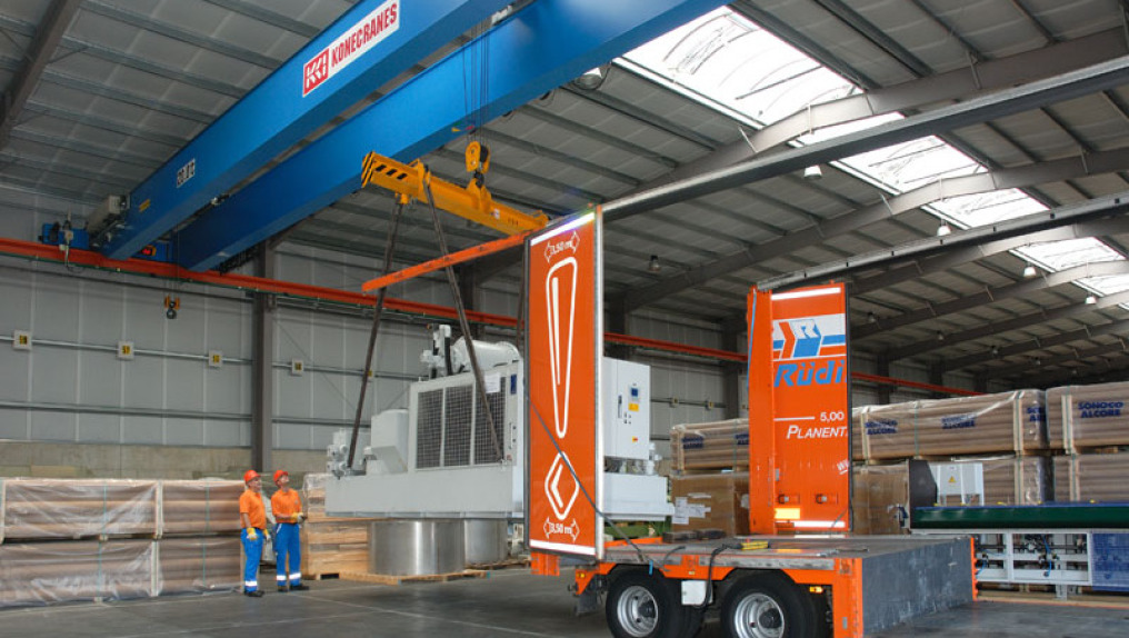 Two employees with helm in orange/blue work with the remote control. A large, gray machine is hanging from a crane and is to be lifted into a completely open truck.