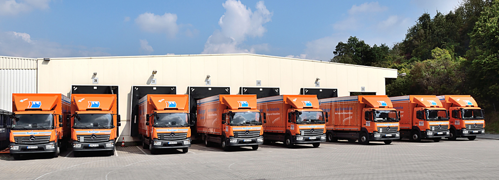 Eight trucks of different sizes are parked in front of five ramps of the transshipment hall. There are trees in the background on the right, the sky is blue with clouds.