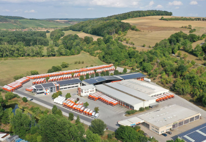 The headquarters of the forwarding company Rüdinger Germany, photographed by a drone. The whole forwarding company is surrounded by nature: fields and wooded areas. You can see buildings and a lot of trucks.