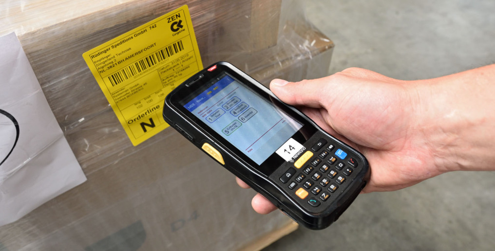 A hand with a hand scanner can be seen in front of a packed box with a yellow label. It is photographed as if the label is being scanned.