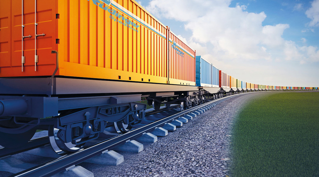 You see an infinitely long train with many colorful containers in a long left curve. On the right side is a green meadow. The leftmost container is huge and orange, and you can see the Rüdinger logo.