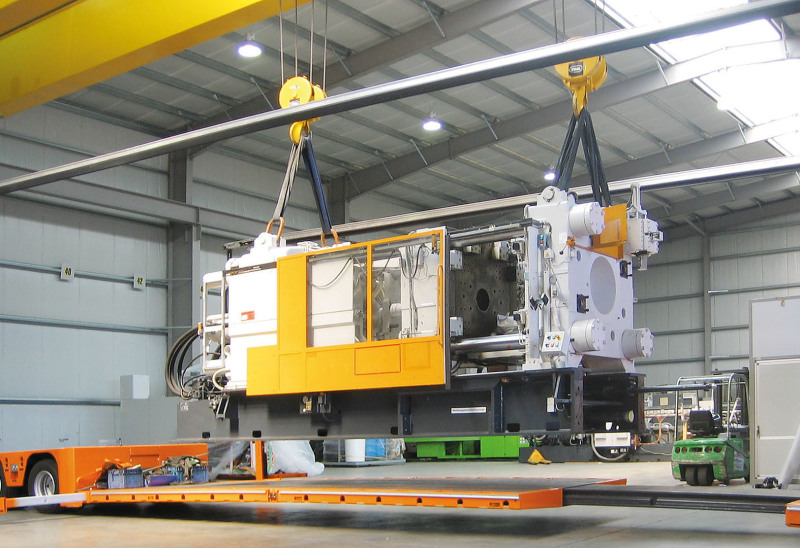A machine in gray and bright yellow hangs from a 32-ton crane. Below it, you can see the orange loading area of a tarpaulin low-loader.