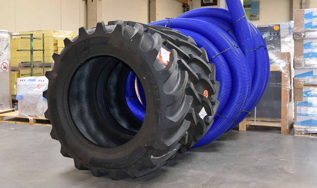 One looks at some packed goods in a warehouse. In front and very dominant are two huge tires that probably belong to a construction site vehicle. Behind them are dark blue tubes wound into several circles.