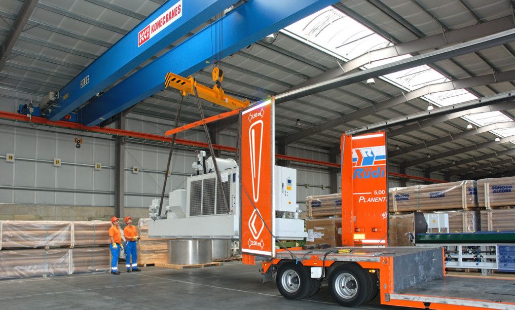 Two employees wearing helmets and blue/orange clothing are working with the remote control and operating a large gray machine on the left side of the picture. On the right is a completely open truck trailer where you can no longer see any tarpaulin.
