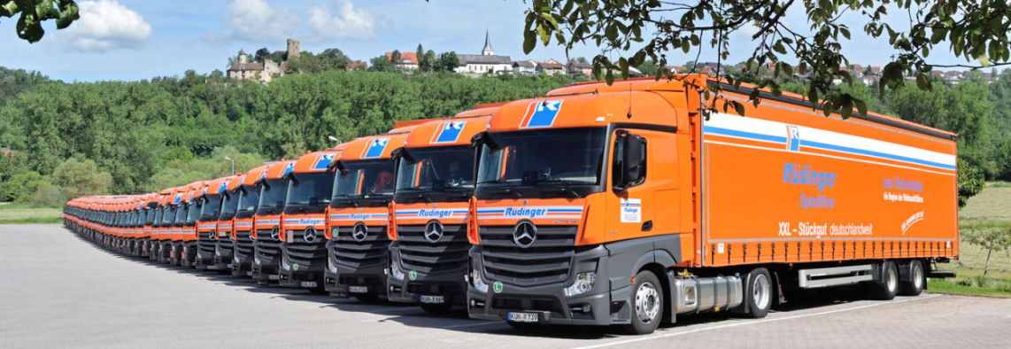 A long row of neatly lined up tarpaulin low-loaders gets smaller and smaller in the distance. Behind the trucks you can see forest, above it the castle and church of Krautheim.