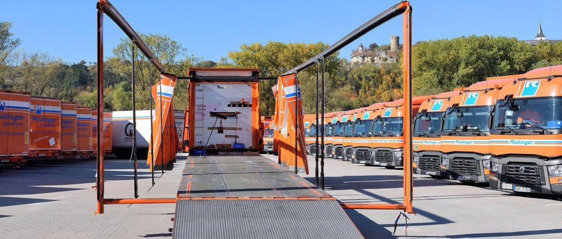 A long row of neatly lined up tarpaulin low-loaders gets smaller and smaller in the distance. Behind the trucks you can see forest, above it the castle and church of Krautheim.