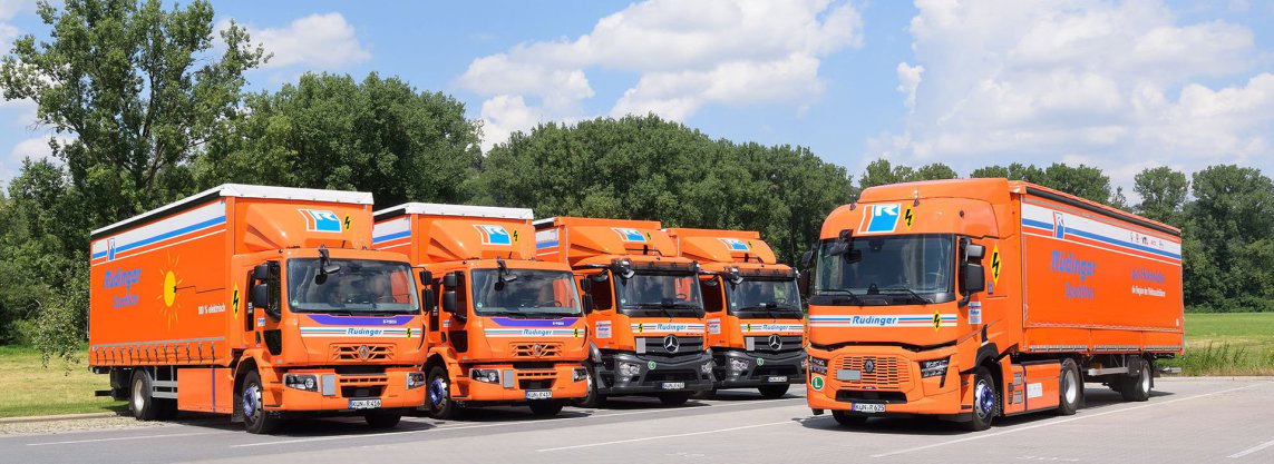 A bright green and yellow agricultural vehicle stands on the lowered dropside, which forms a flat ramp surface. On the left, you can see the trailer of a truck open on the side. In the background, more freight forwarding is visible.
