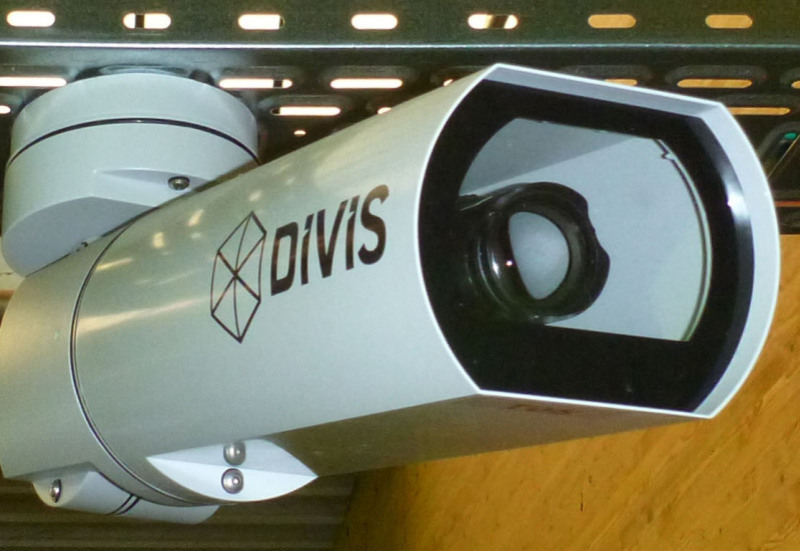 A large camera of the DIVIS video system hangs on the ceiling in front of a yellow wall. You can see the camera at an angle from the side.