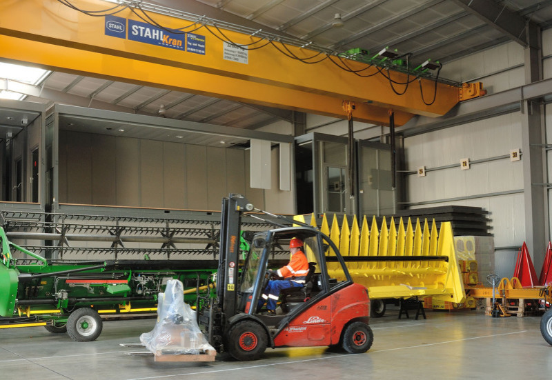 In front of a large 32-ton crane on the warehouse ceiling is a light green and a yellow agricultural machinery. In front of it, a forklift moves from right to left and has loaded a pallet with a small good.