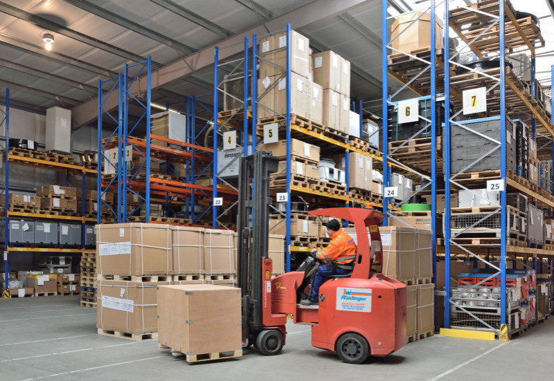 In a newly built warehouse you can see many shelves all well filled with different goods. In the front drives a forklift, which transports a wooden box.