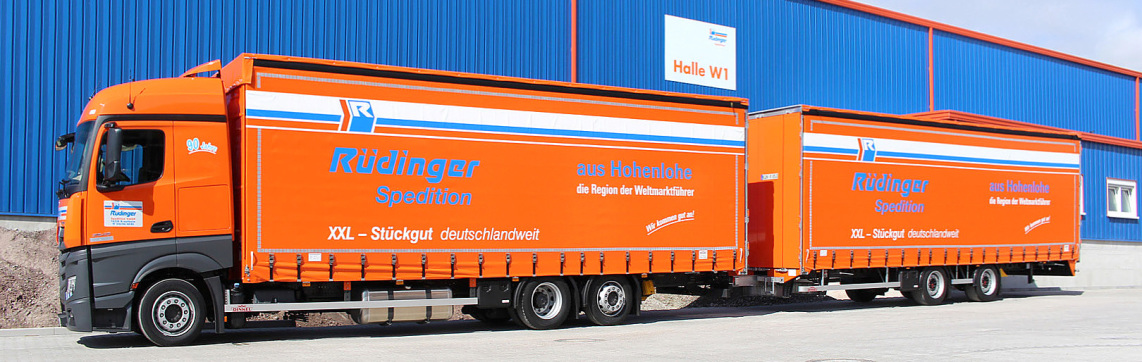 A special long truck with trailer, a so-called giga liner stands in front of a newly built warehouse in the colors blue and orange. You can see it from the side.
