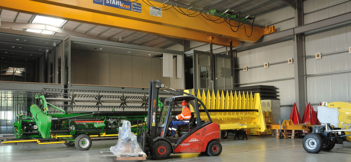 At the top of the hall ceiling is a yellow 32-ton crane. On the storage area are two large agricultural machines, one in yellow, the other in green. In front of them, an employee in a helmet is riding on a forklift and has loaded a small piece of goods.