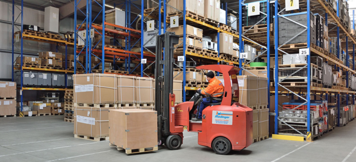 You look sideways at many shelves in a warehouse. All shelves are filled with many different goods in different colors. In the foreground, an employee is riding a special forklift and has loaded a wooden crate.