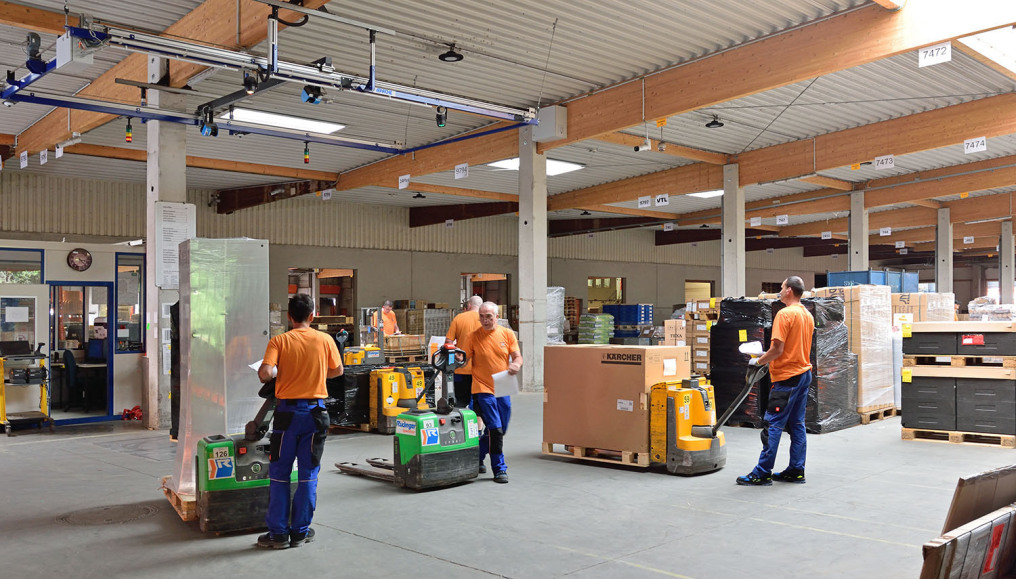 Many employees in orange/blue dress give the impression of hectic activity around the "Apache" measuring and weighing system. Some have goods on their lift trucks.