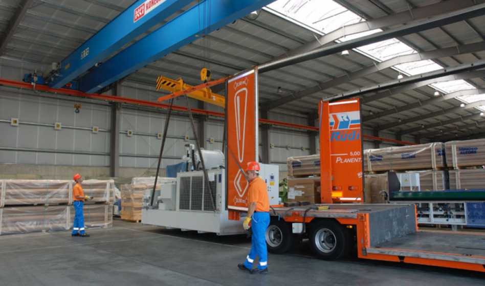 Two employees with orange/blue clothing and a helmet on lift a large gray machine by remote control. On the right you can see a completely opened tarpaulin low loader. You can see only the steel skeleton of the trailer.
