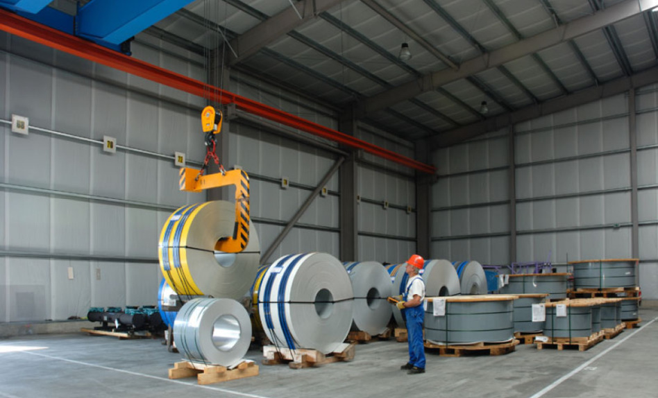You look into the back corner of a warehouse. A man with a helmet is lifting a huge coil of metal with a smaller crane. More such coils are on the floor.